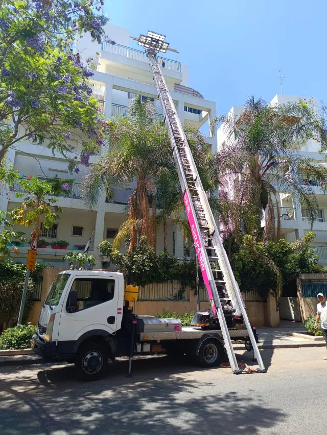 משאית המנוף עם הזרוע הוורודה פרוסה לגובה מול בניין מגורים